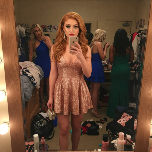 A woman in a sparkly rose gold dress takes a mirror selfie. Three other women in dresses are visible in the background of the messy room, getting ready.